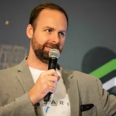 Ryan Selkis, a speaker with a beard, holds a microphone and wears a grey blazer over a white t-shirt.
