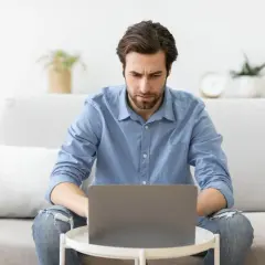 A man with a beard, wearing a blue shirt and ripped jeans, looks intently at a laptop on a small table.