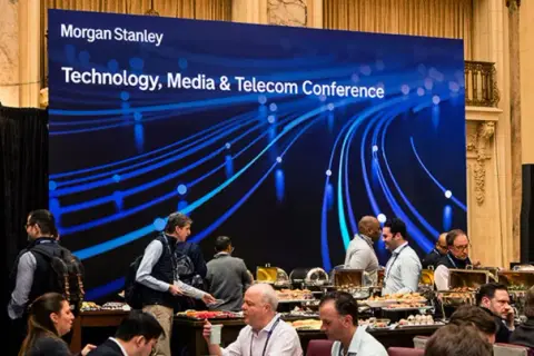 Attendees mingle at a Morgan Stanley TMT conference, with a large screen displaying the event title and a blue, abstract network graphic.