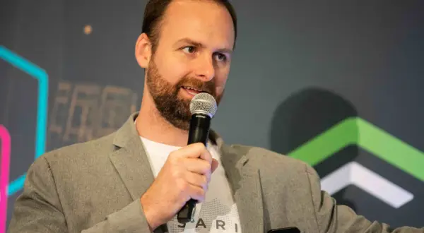 Ryan Selkis, a speaker with a beard, holds a microphone and wears a grey blazer over a white t-shirt.