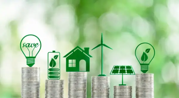 Stacks of coins topped with green icons of a lightbulb, battery, house, wind turbine, and solar panel, illustrating how to invest in renewable energy.