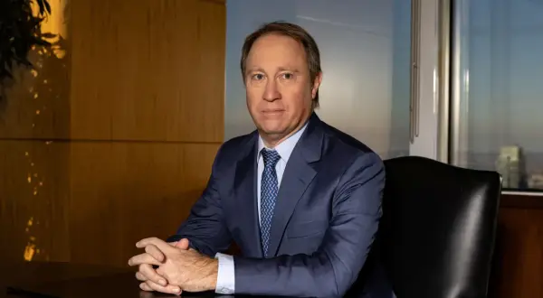A man in a suit sits at a desk, hands clasped, in an office with a city view. He looks like he's discussing important Morgan Stanley acquisitions.