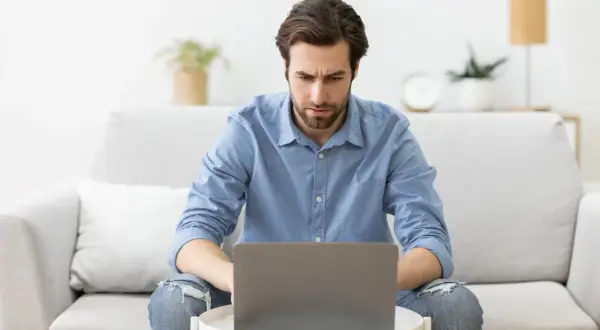 A man with a beard, wearing a blue shirt and ripped jeans, looks intently at a laptop on a small table.