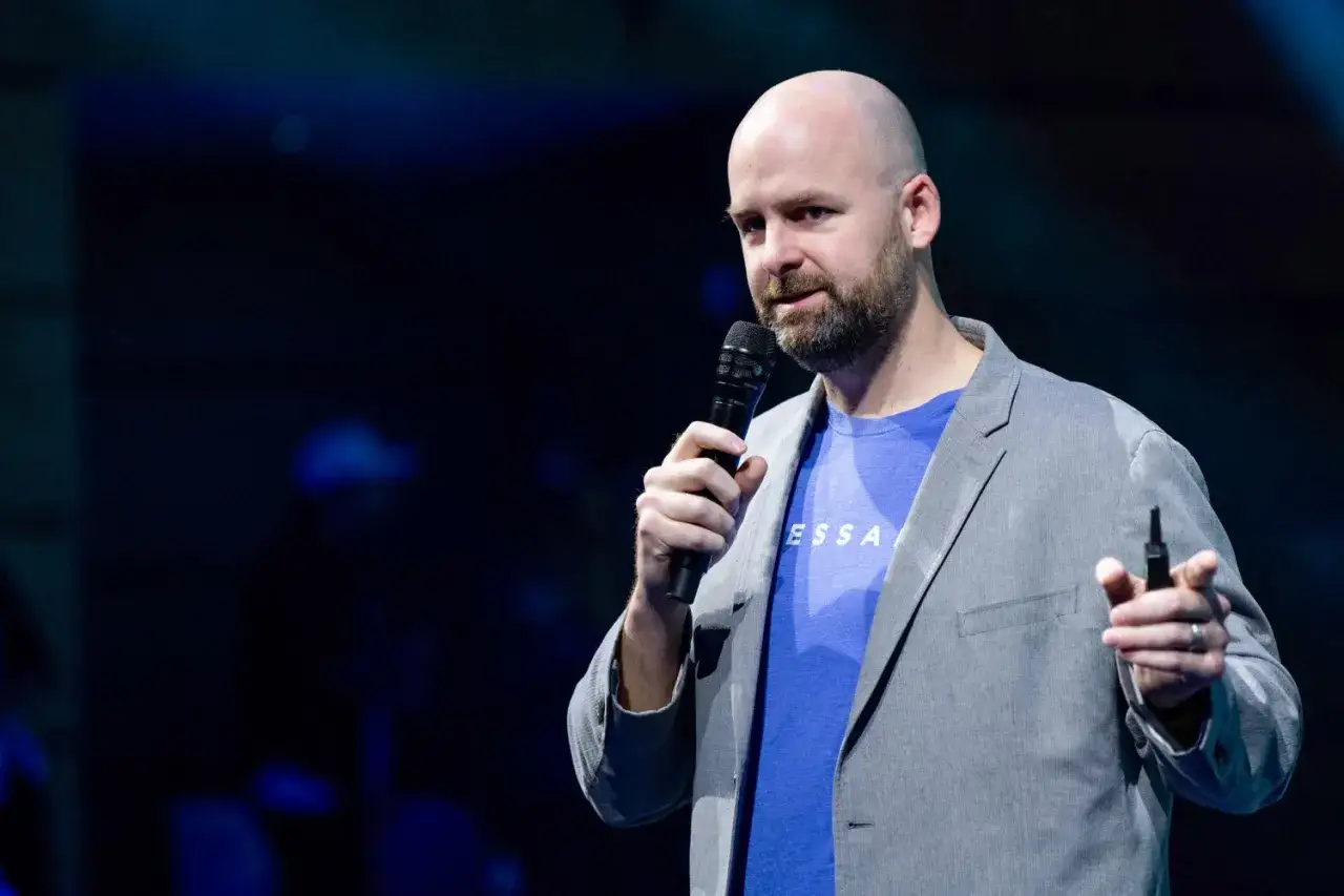 Ryan Selkis, a bald man with a beard, speaks into a microphone on a dark stage, wearing a grey blazer over a blue t-shirt.