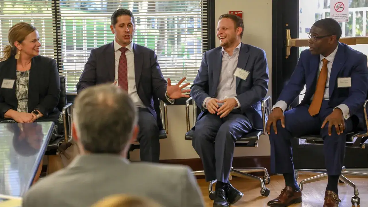 Panelists discuss alternative investment partners. Four professionals in suits sit in chairs, engaged in conversation.
