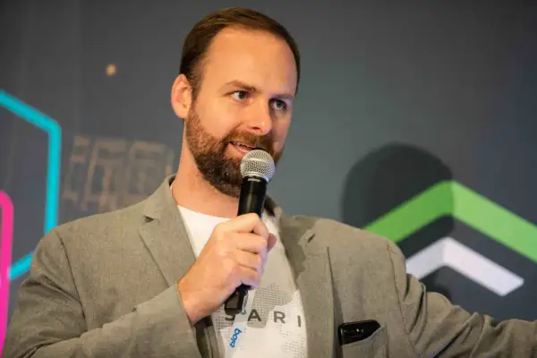 Ryan Selkis, a speaker with a beard, holds a microphone and wears a grey blazer over a white t-shirt.